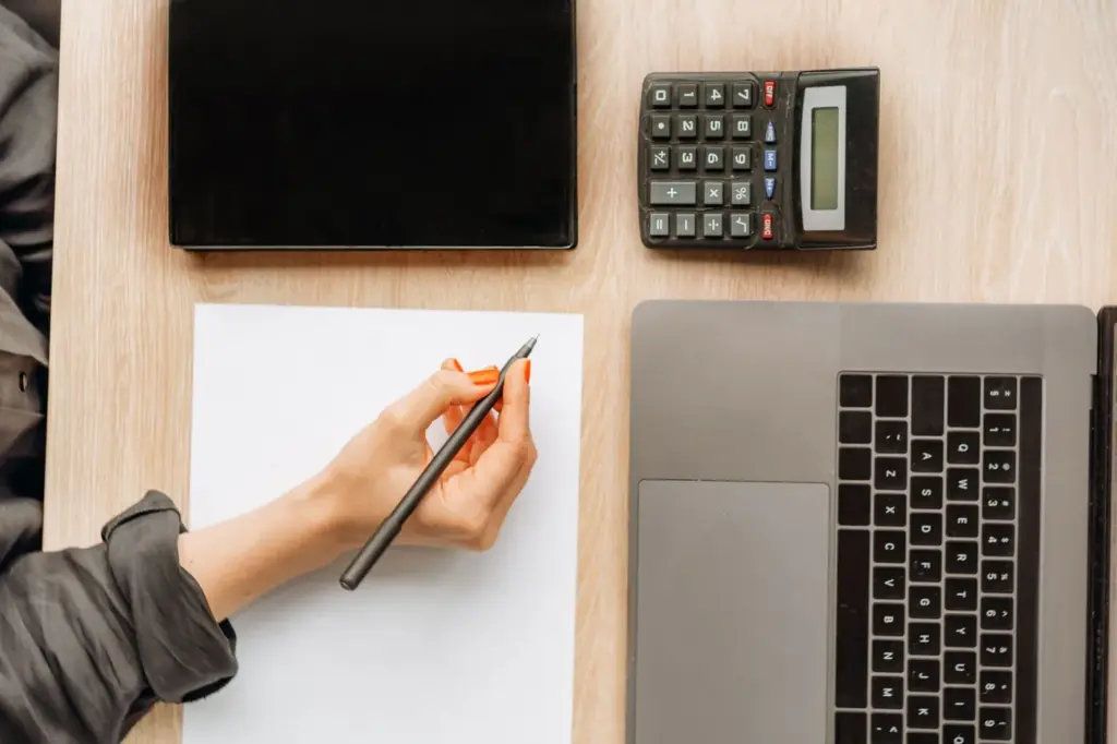 Overhead view of a hand holding a pencil over a blank sheet of paper beside a calculator and laptop