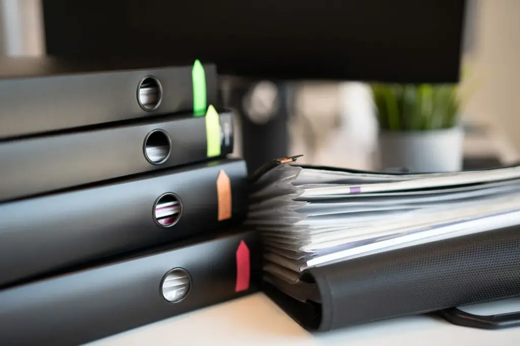 A stack of ring binders and documents with coloured tabs on a desk
